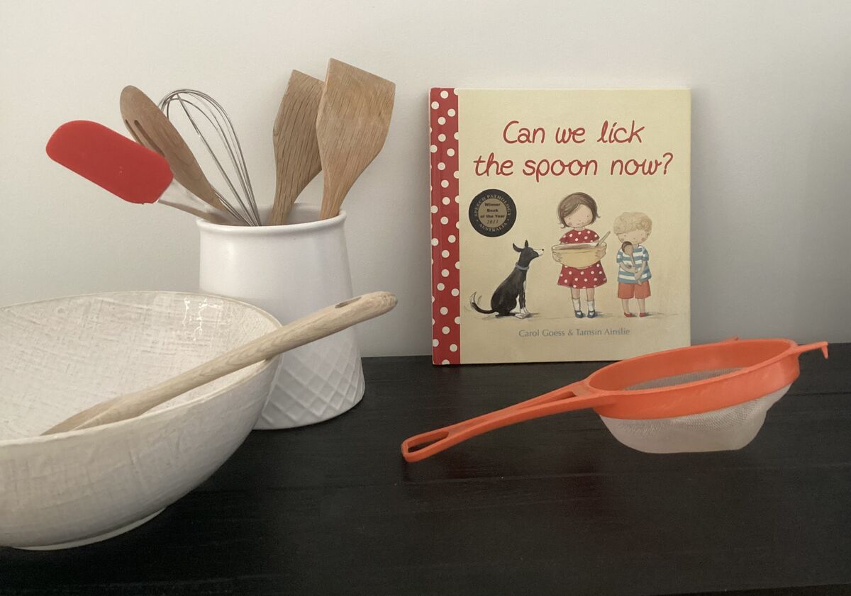 Can We Lick the Spoon Now? picture book propped up alongside a mixing bowl, wooden spoons, a whisk, and a red sieve on a dark surface